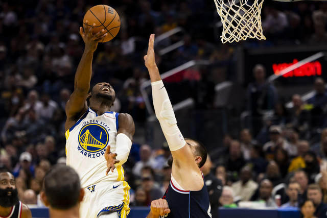 A Golden State Warriors player leaps for a layup as a defender with an outstretched arm tries to block the shot near the basketball hoop.